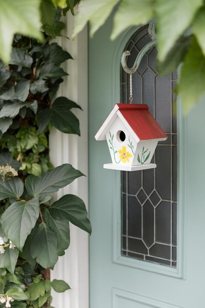 Colorful birdhouse with red roof and floral design hanging by a teal door, surrounded by green leaves.
