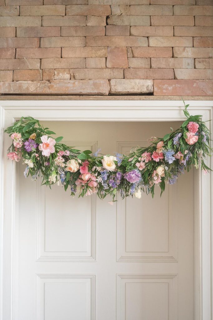 Floral garland with pink, purple, and white flowers hanging on a door against a brick wall background.