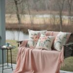 Cozy porch swing with floral pillows and pink blanket, next to a table with a teacup and book, overlooking a serene forest.