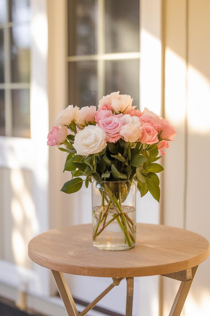 Pink and white roses in a glass vase on a round wooden table, indoors with soft lighting.